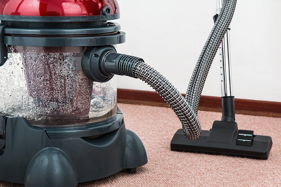 A yellow canister vacuum cleaner with a black hose and a floor attachment is positioned on a patterned area rug with floral motifs in a room with hardwood flooring. Adjacent to the rug, there is a woo