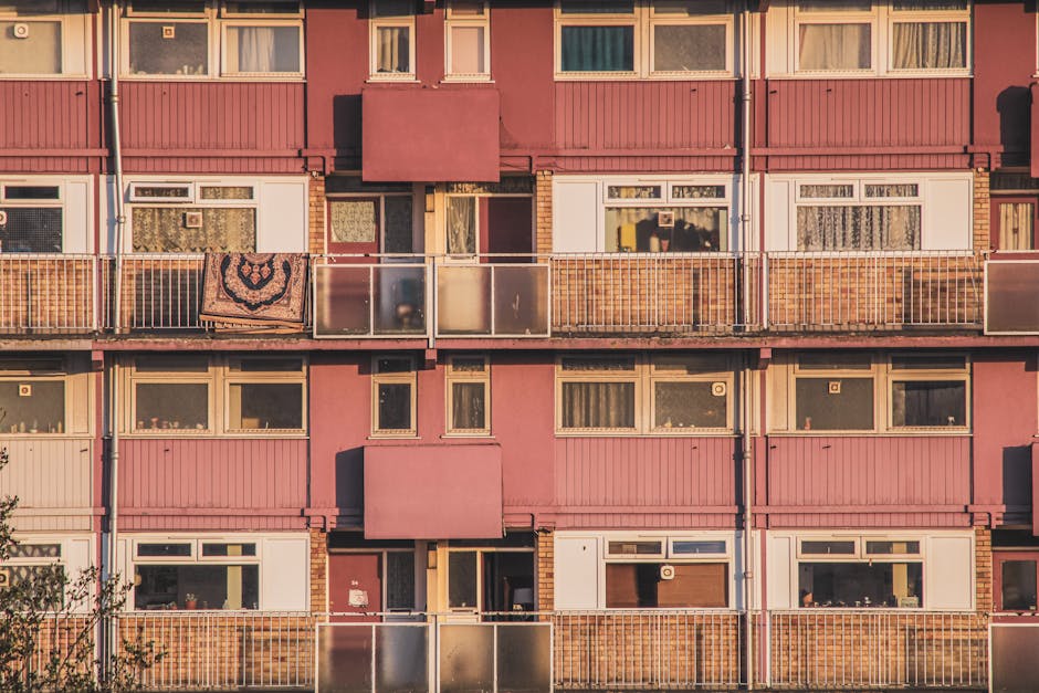 A multi-story residential apartment building featuring a pink facade with white and beige window frames. The balconies have transparent glass panels with metal railings, some with items such as a patt