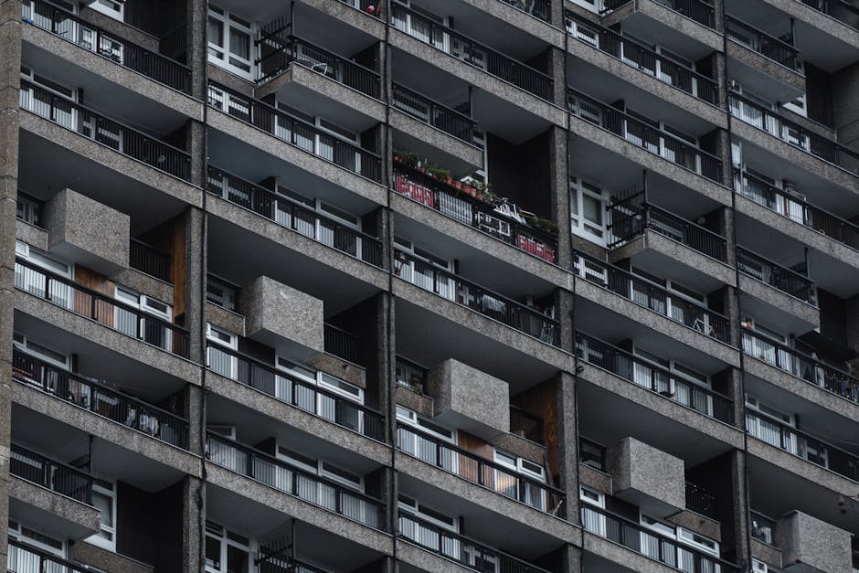 A multi-story residential apartment building featuring a pink facade with white and beige window frames. The balconies have transparent glass panels with metal railings, some with items such as a patt